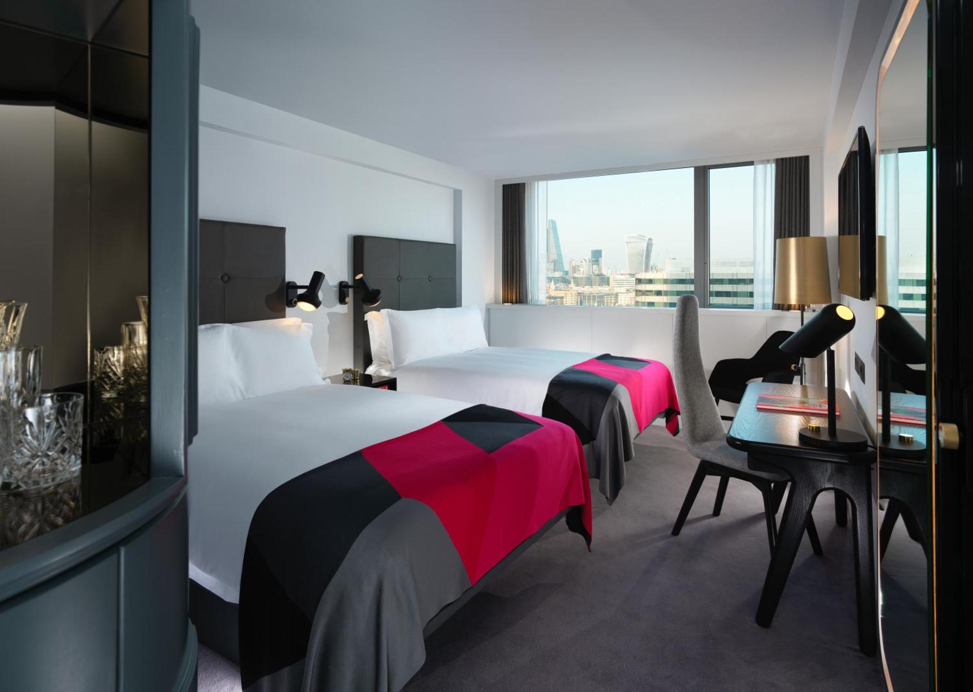 Готель Sea Containers Лондон Екстер'єр фото The photo shows a modern hotel room featuring two beds with colorful throws in shades of pink and gray. The room has large windows that provide a view of the outside, and there are sleek, contemporary furnishings including a desk and chair. The walls