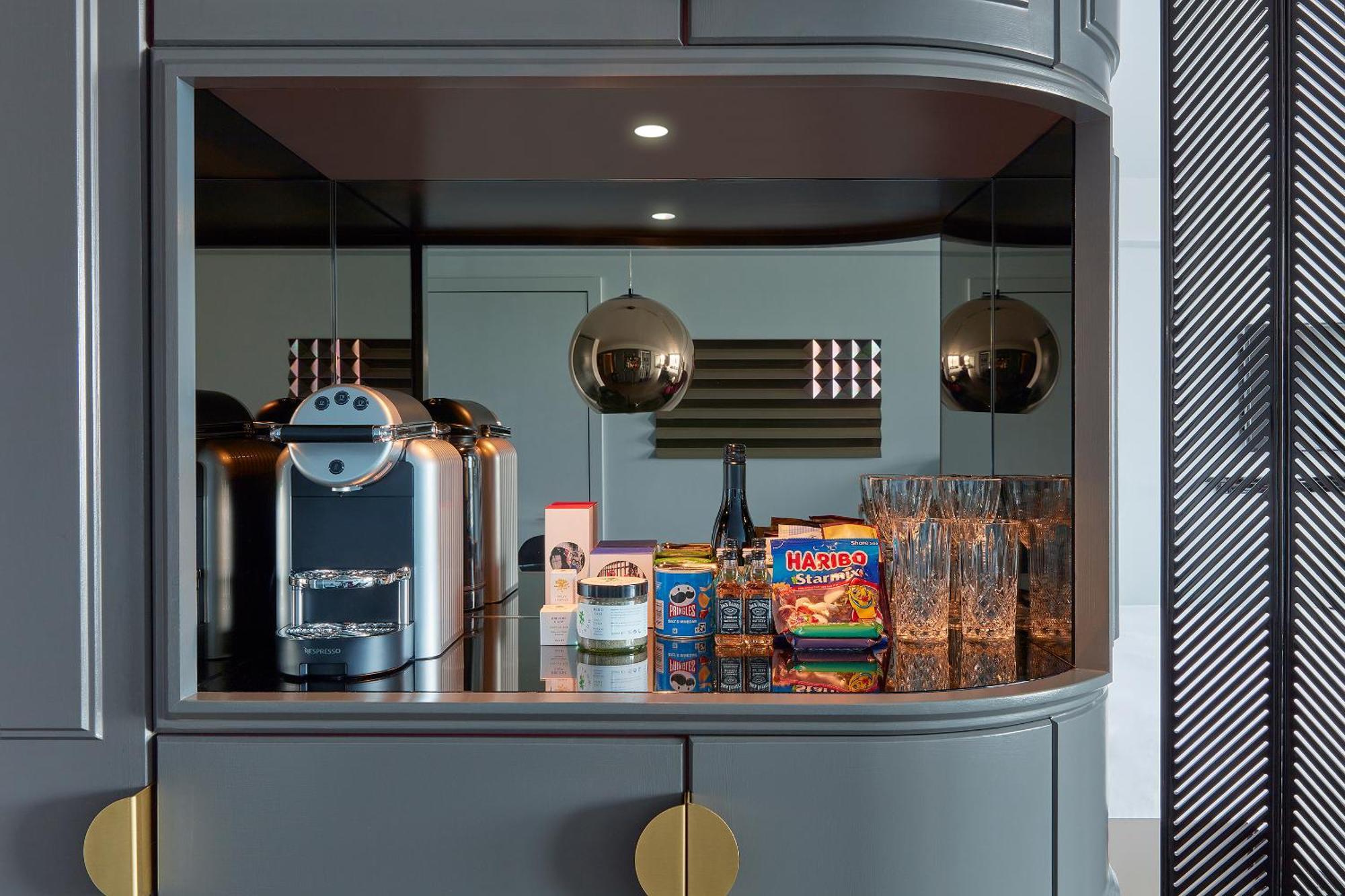 Готель Sea Containers Лондон Екстер'єр фото The photo displays a modern kitchen nook with a sleek design. It features a coffee machine on the left, placed on a dark countertop. Next to it, there are several glass drinking glasses arranged neatly. Prominently displayed is a colorful box of snac