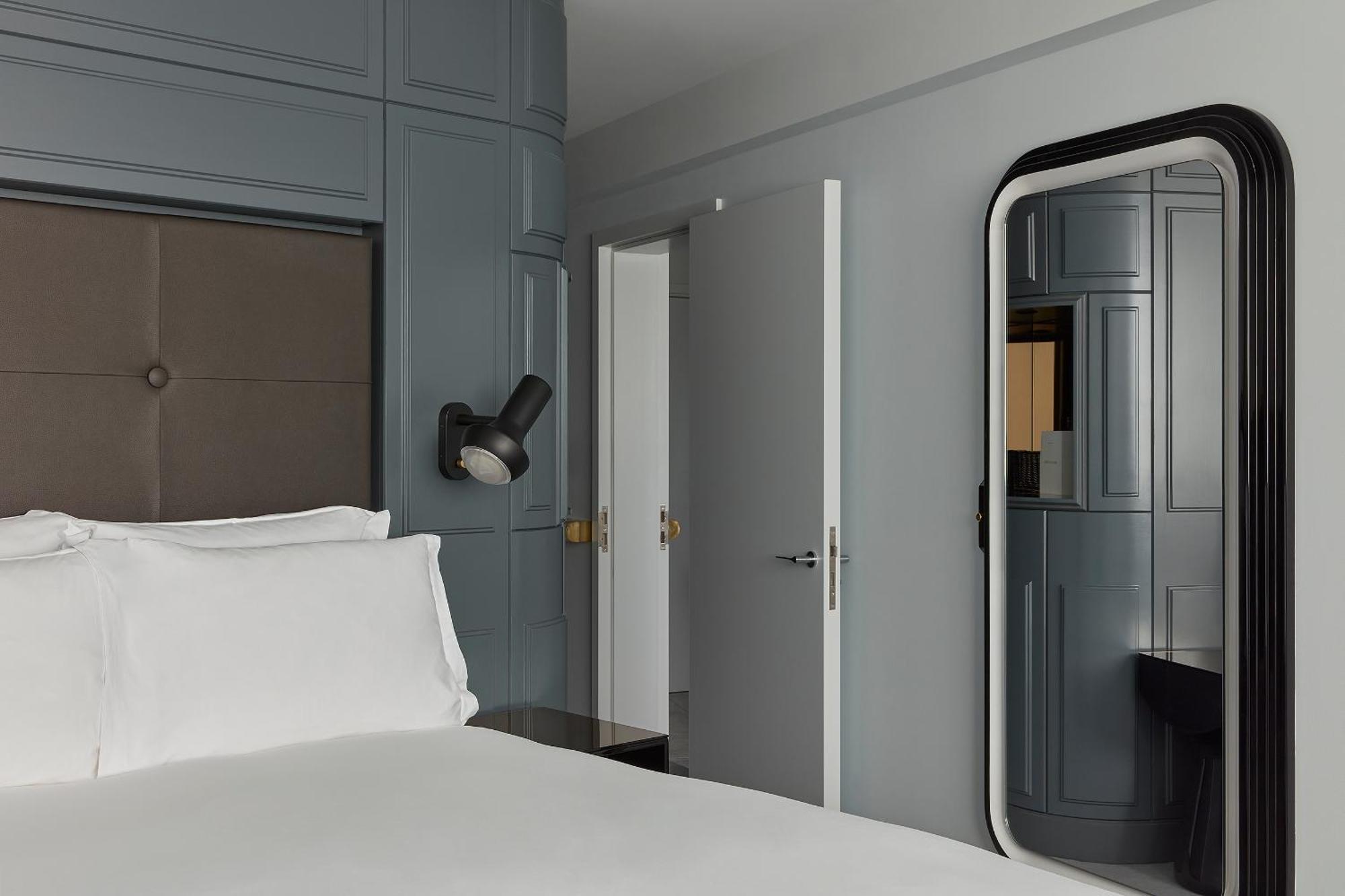 Готель Sea Containers Лондон Екстер'єр фото The photo shows a modern hotel room or bedroom with a minimalist design. There is a large bed with white linens positioned prominently in the foreground. The walls are painted a muted gray, and some sections have textured paneling. To the right, ther