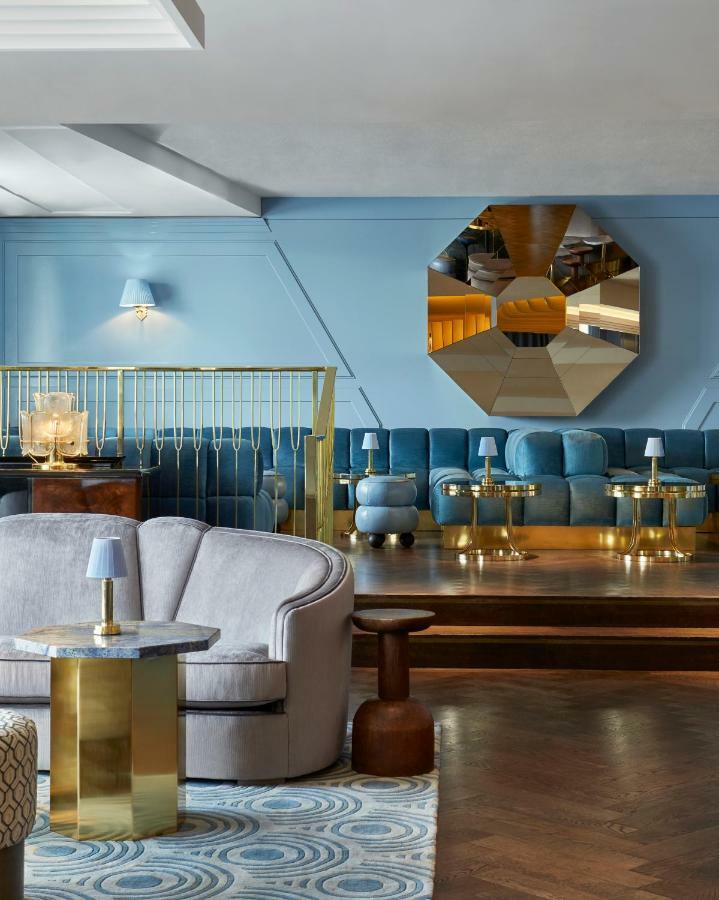 Готель Sea Containers Лондон Екстер'єр фото The photo depicts a stylish lounge area featuring a soft blue color palette. There is a plush sofa in light blue and a series of small tables with brass legs scattered throughout the space. A large, faceted mirror is mounted on the wall, adding a mod